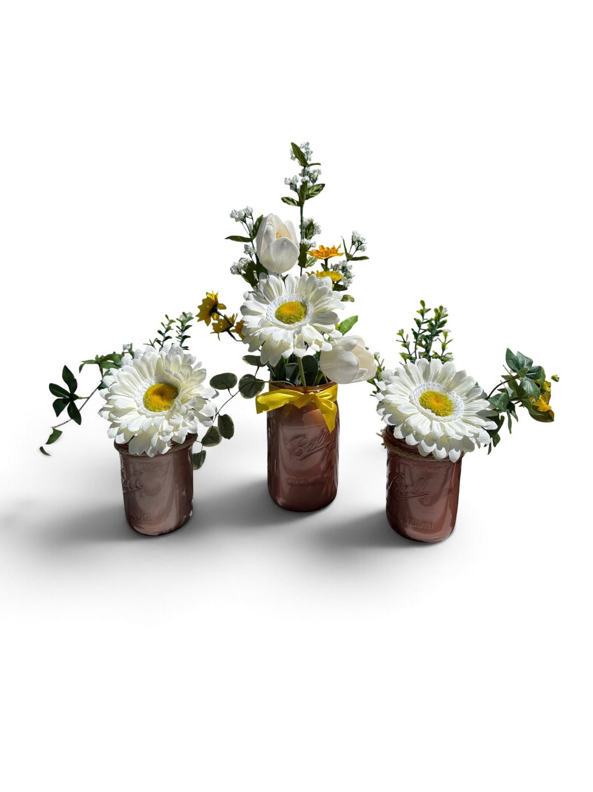 Three small copper-colored flower pots arranged together on a white background, each filled with white daisies, green foliage, and small yellow wildflowers. The center pot has a yellow ribbon tied around the neck.