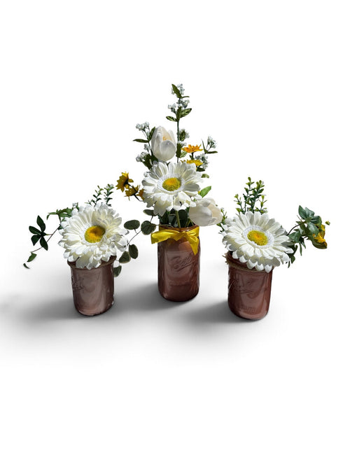 Three small copper-colored flower pots arranged together on a white background, each filled with white daisies, green foliage, and small yellow wildflowers. The center pot has a yellow ribbon tied around the neck.