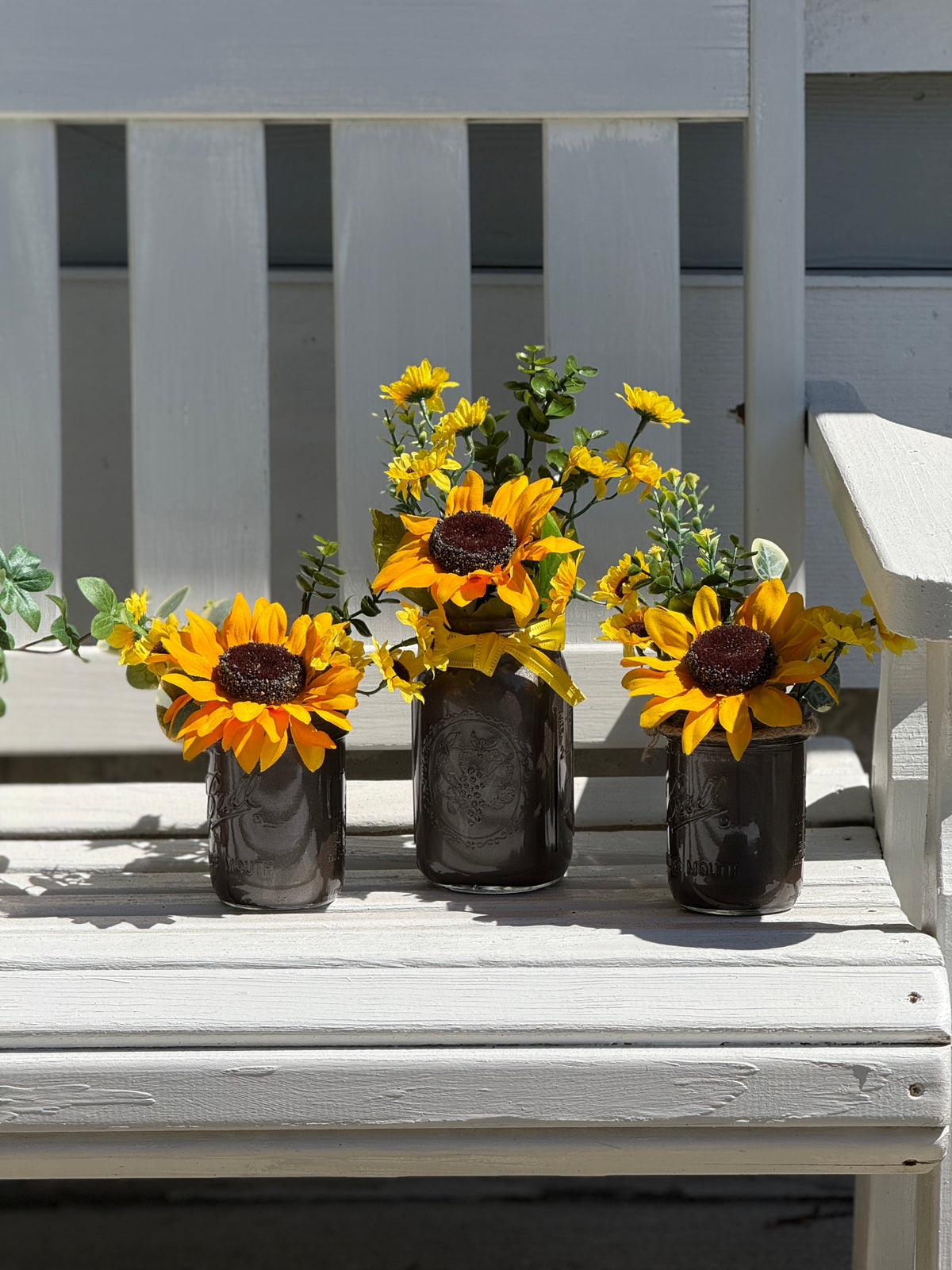 Barking Gardener farmhouse faux sunflower arrangement in three rustic brown mason jars on a white wooden bench.
